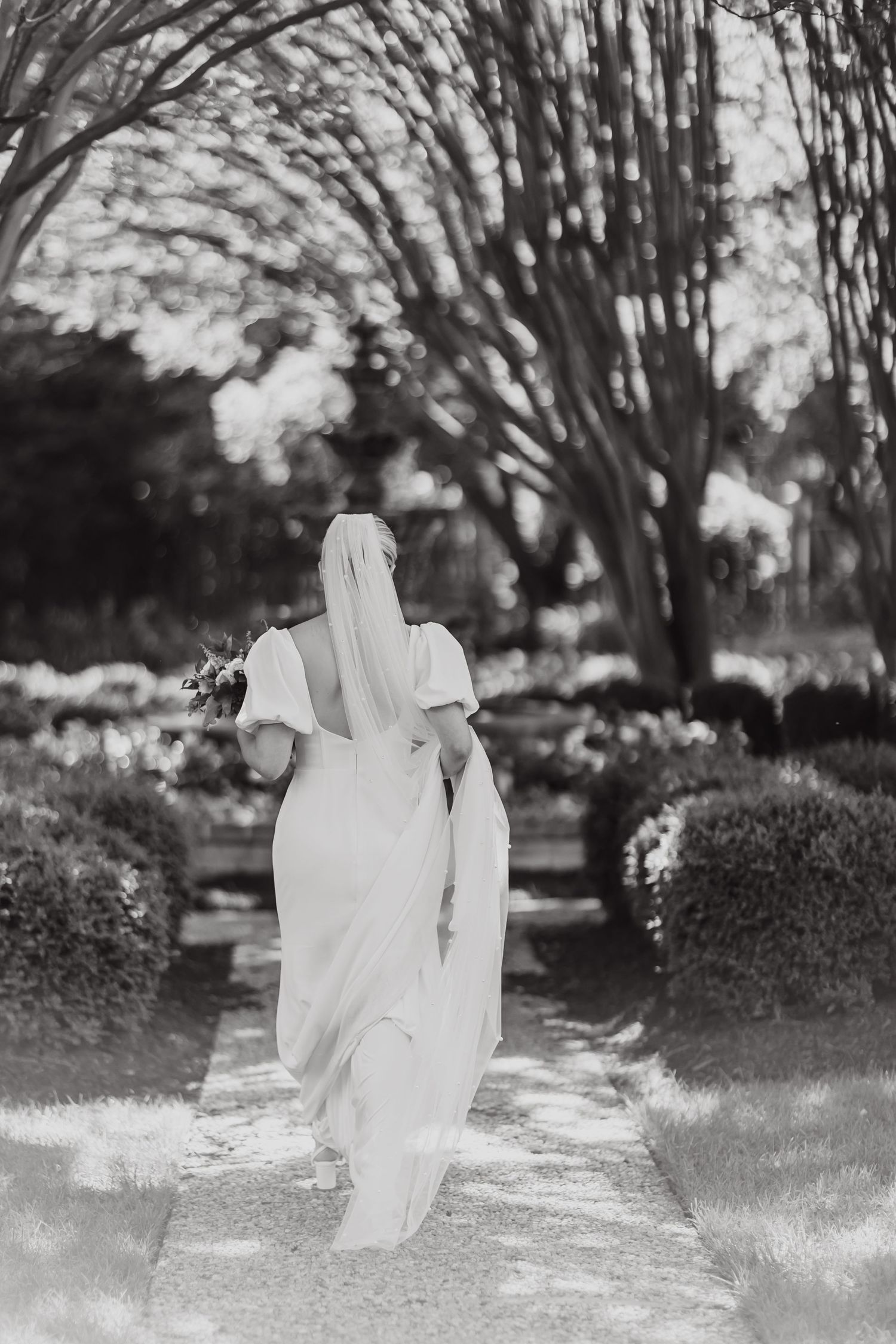 Wedding couple walking through garden path with overhanging trees creating natural archway.