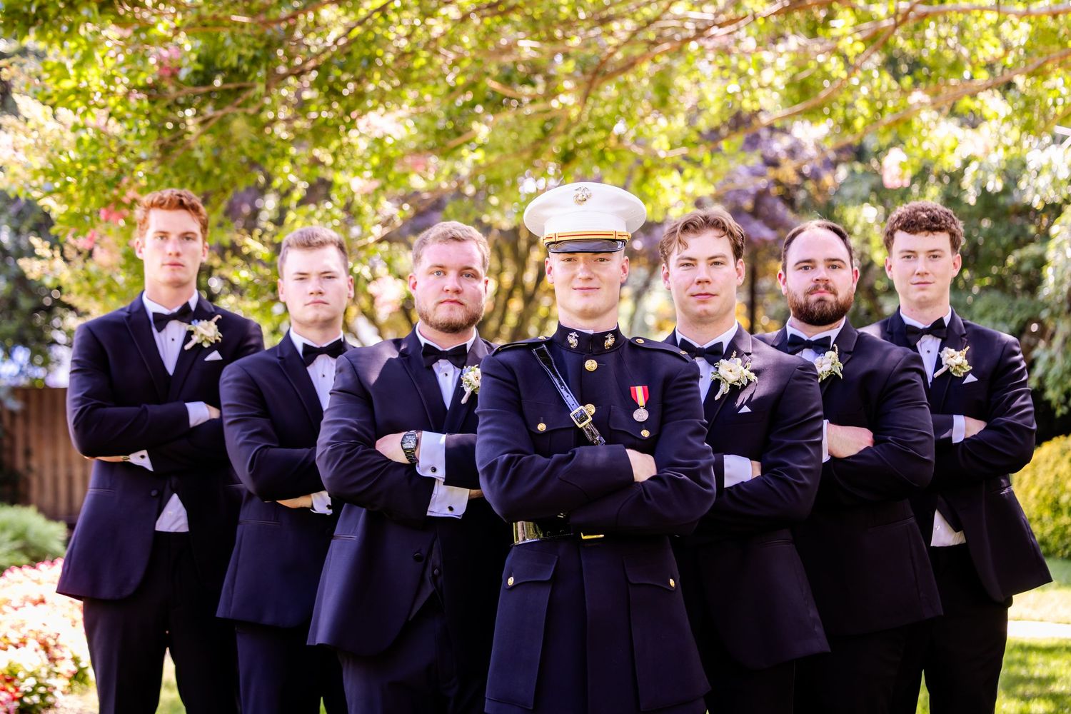 Groomsmen in navy uniforms stand in formation for a formal wedding photo outdoors.