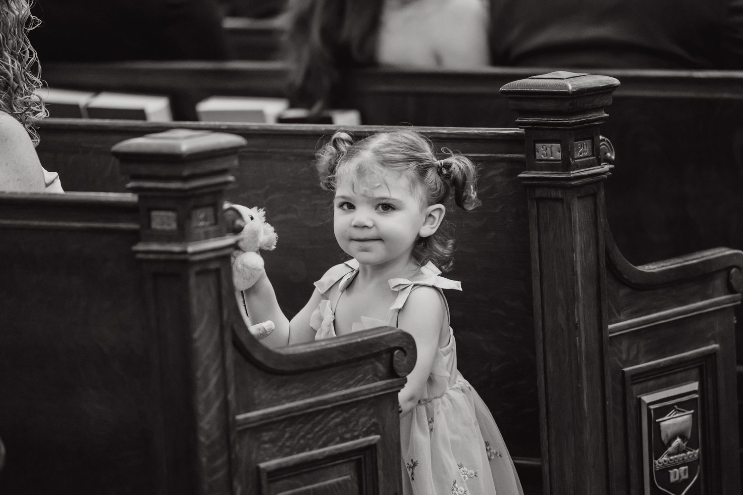 Young child in a dress stands at a church pew in black and white vintage photograph.