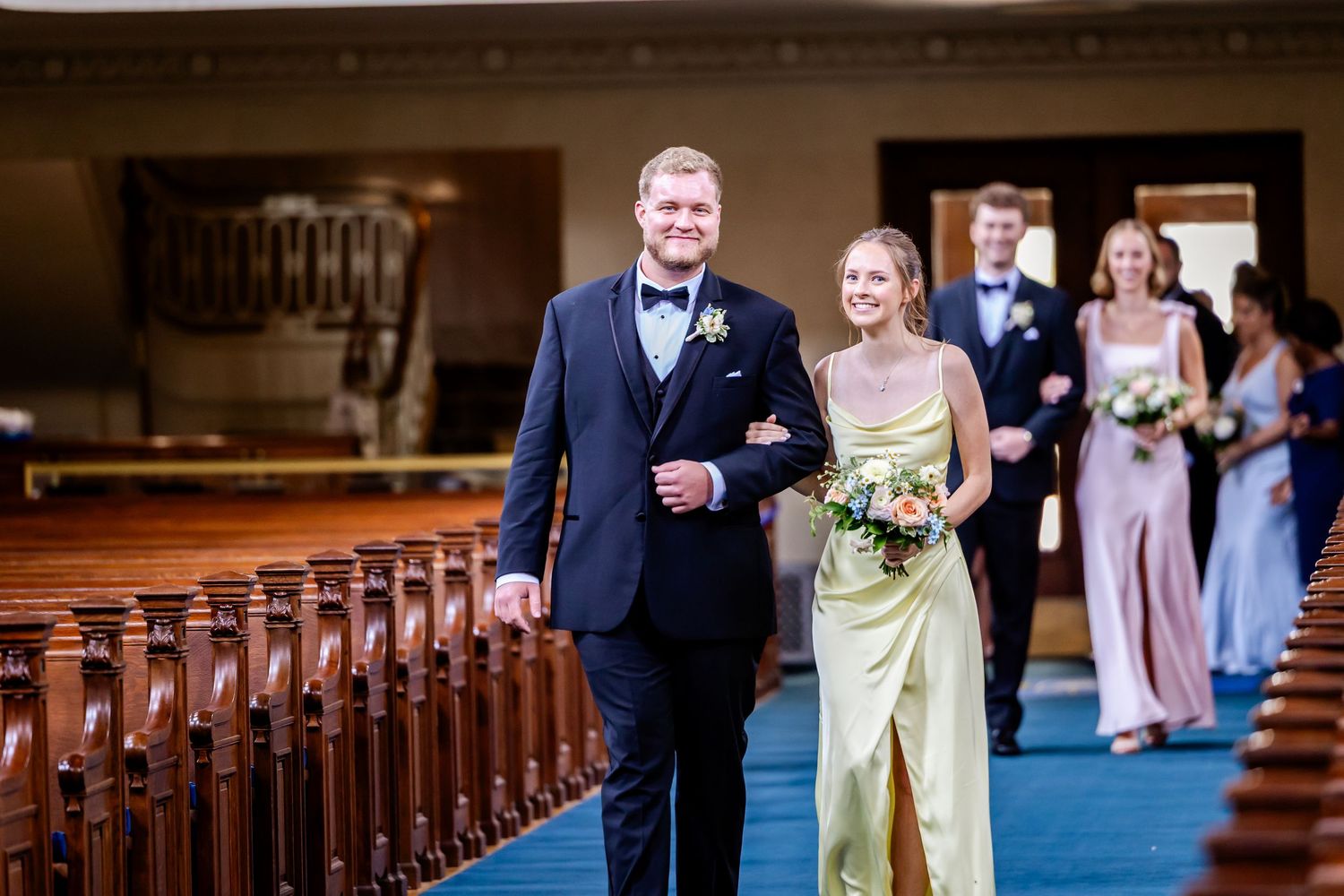 Wedding party processes down church aisle with wooden pews and blue carpet.
