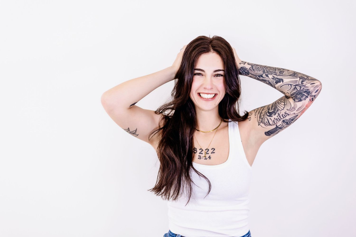 Smiling brunette woman with tattoos on arms wearing white tank top, hands in hair, white background.