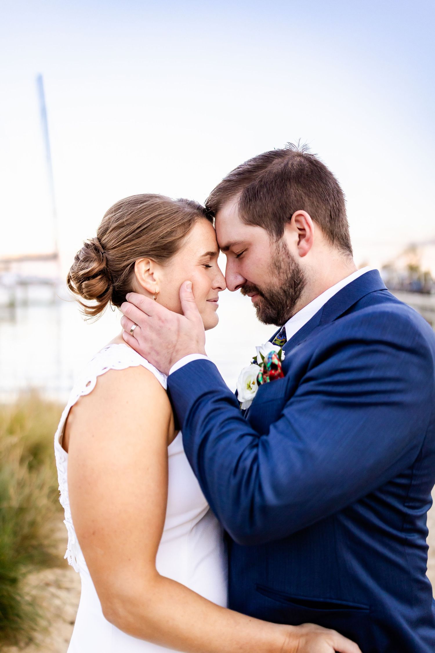 A bride and groom share a tender forehead touch, the groom in a navy suit with boutonniere, near a waterfront.