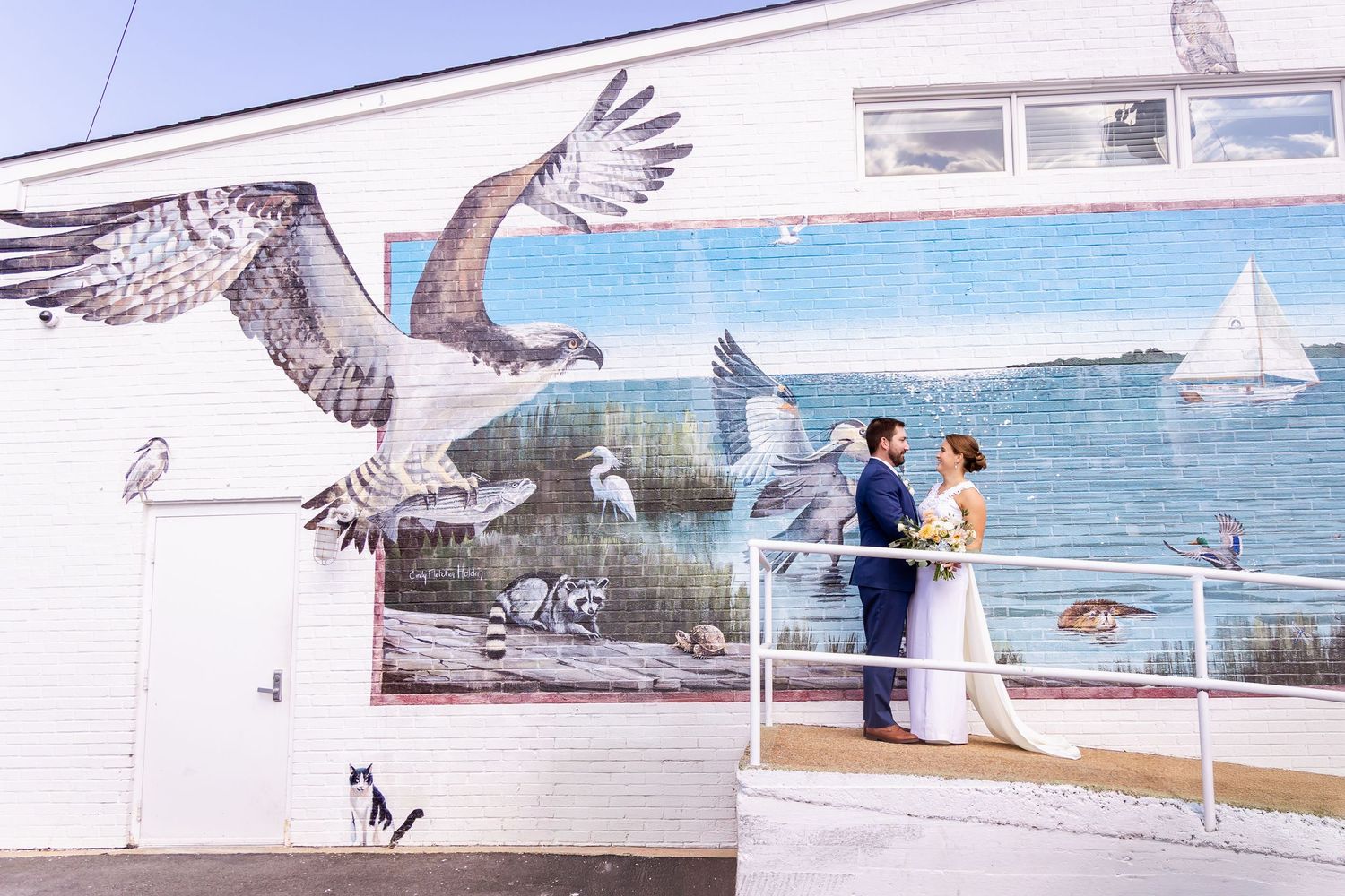 Bride and groom share a romantic moment in front of a colorful coastal mural with birds and sailboat.