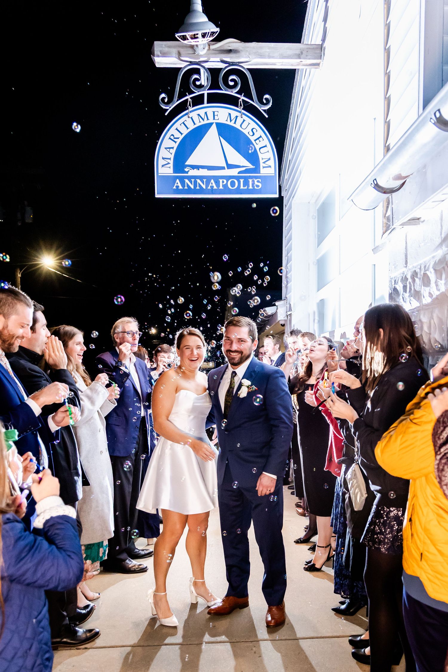 Bride and groom exit Maritime Museum Annapolis amid bubbles and cheering guests at night wedding celebration.