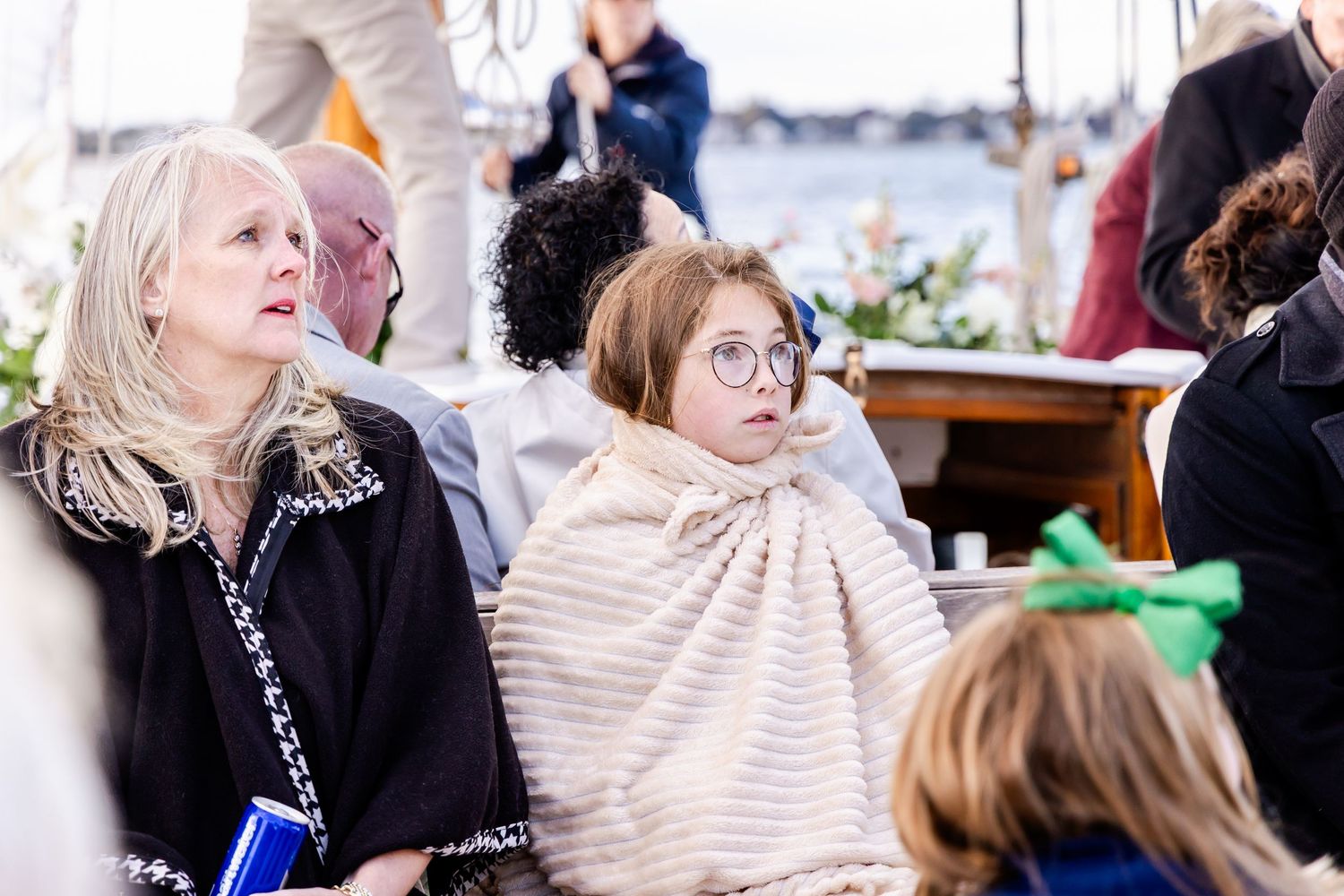 People seated outdoors at a waterfront event, woman with glasses wrapped in striped blanket beside blonde woman in black jacket.