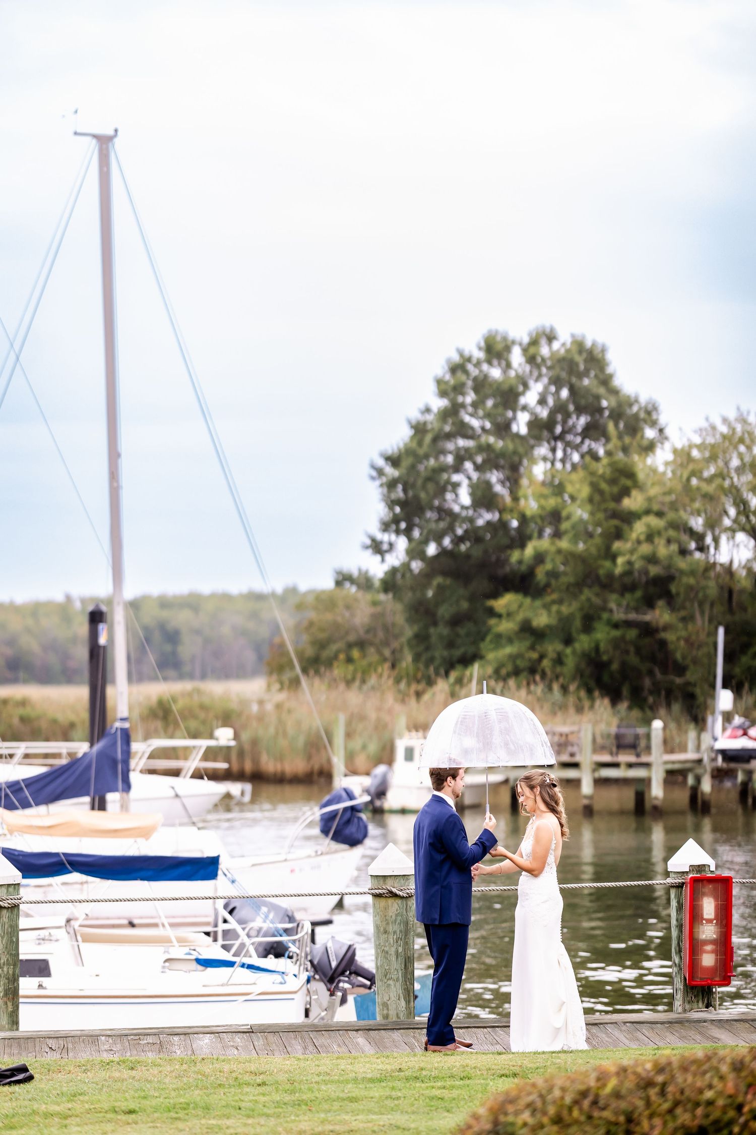A wedding couple poses near boats at a marina with water and trees in the background.