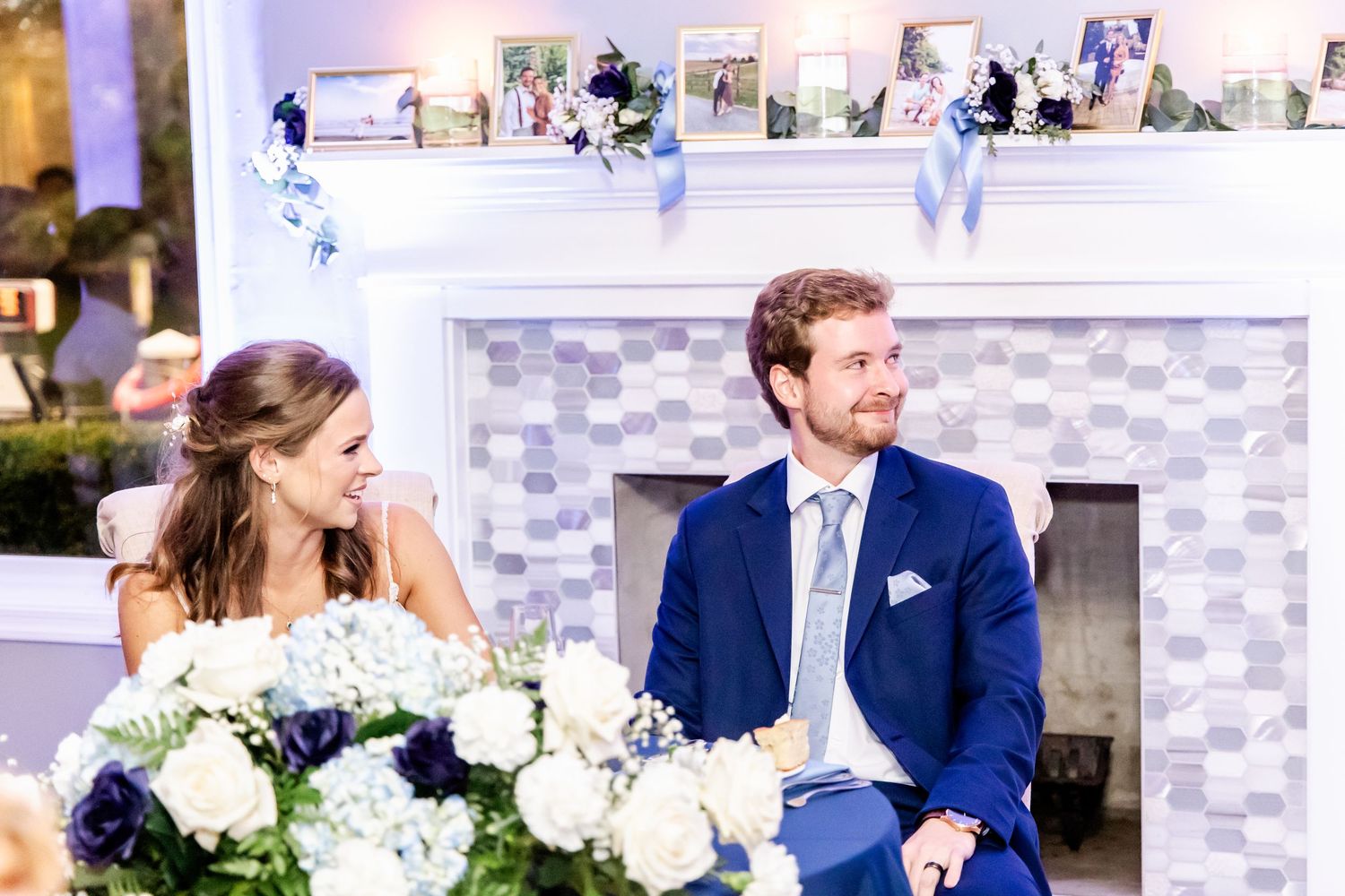 Newlyweds seated by an elegant fireplace with white floral arrangements during their reception.