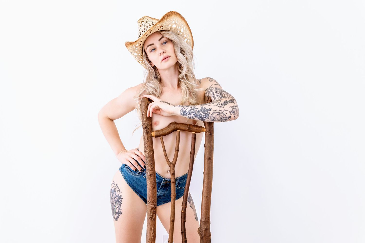 A person in western attire with tattoos poses with a wooden chair against a white background.