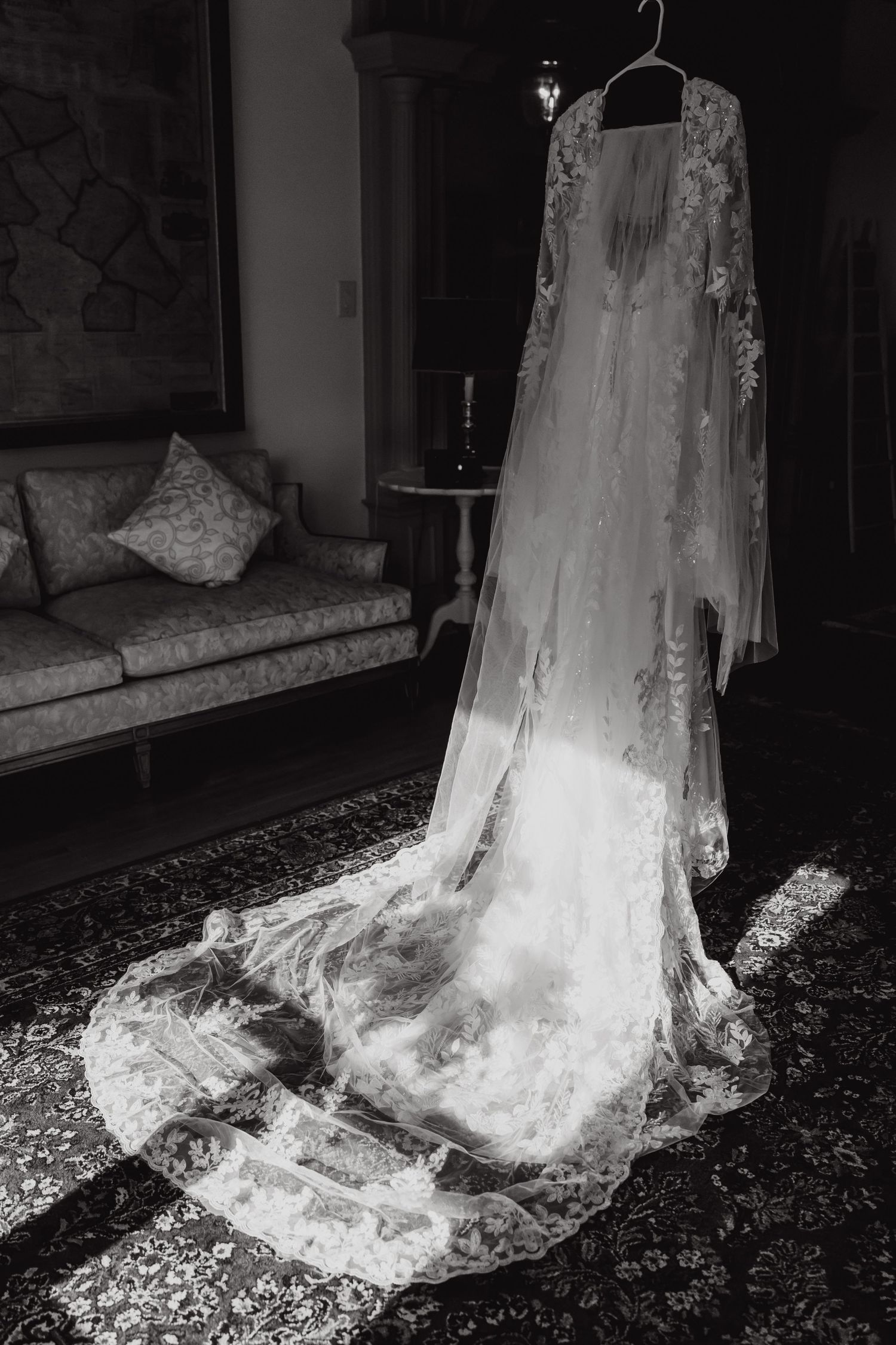 Delicate white lace wedding dress with long train displayed on a hanger in dramatic lighting.