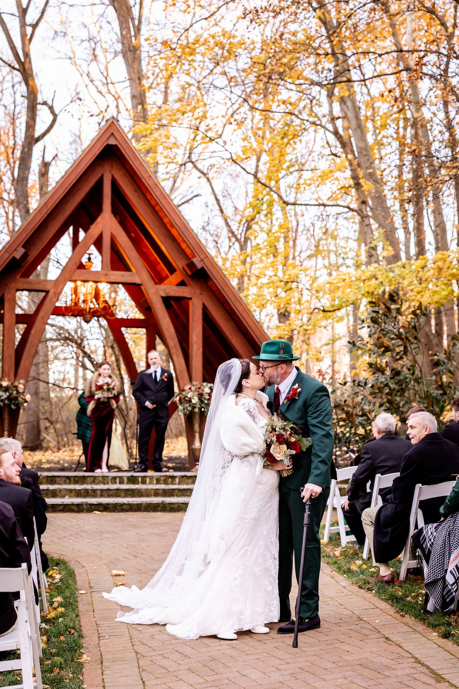 Outdoor wedding ceremony procession under wooden pavilion surrounded by autumn trees.