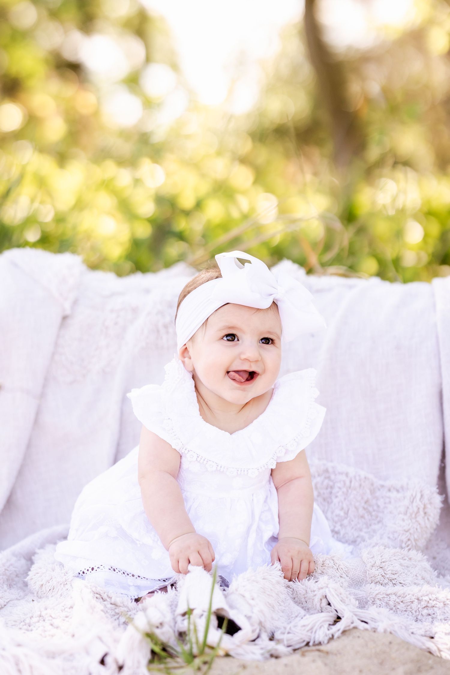 Baby in white dress and bow crawls on lace blanket in sunny outdoor setting with soft bokeh background.