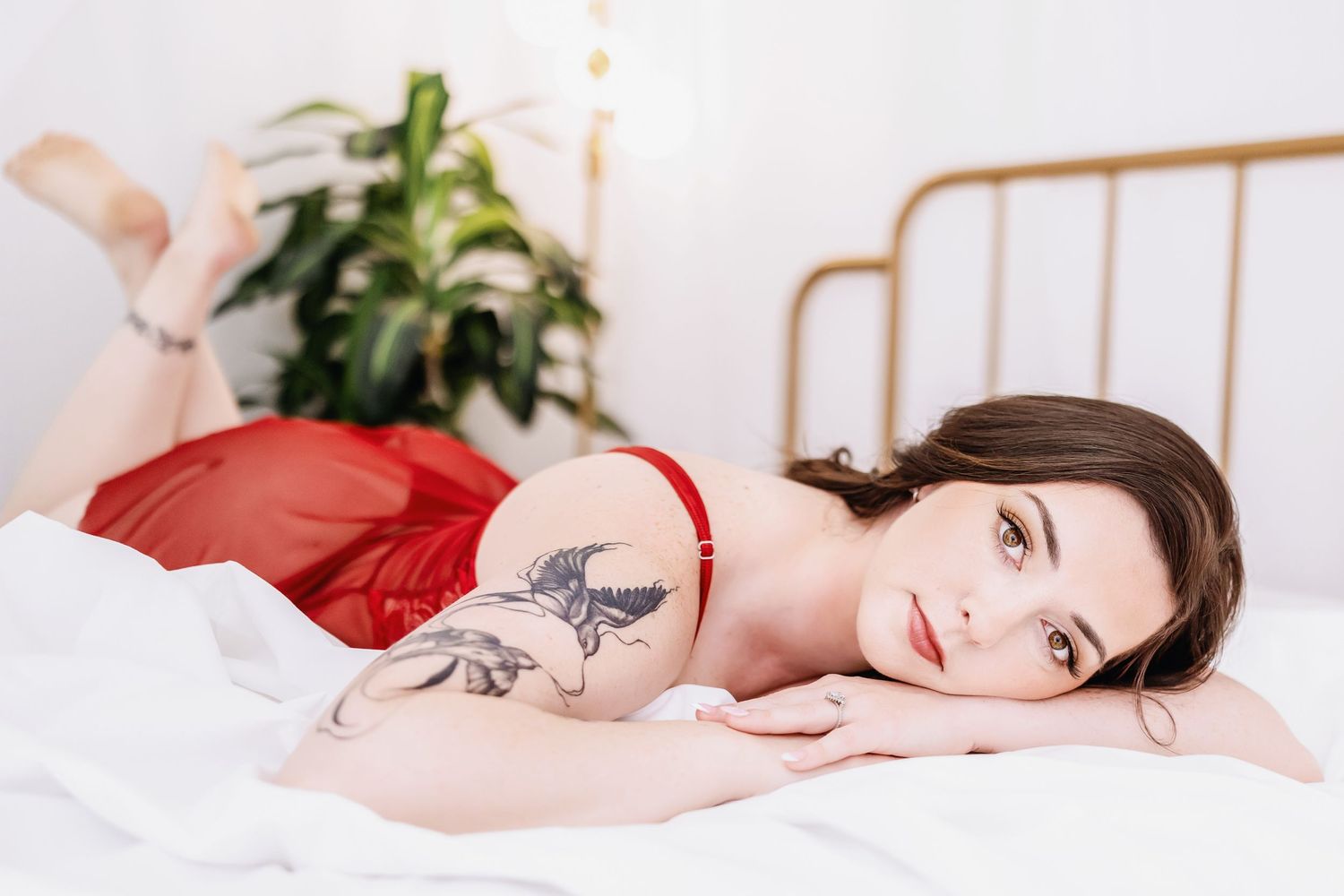 Woman in white bedding with red lingerie and tattoo poses sensually on white sheets.