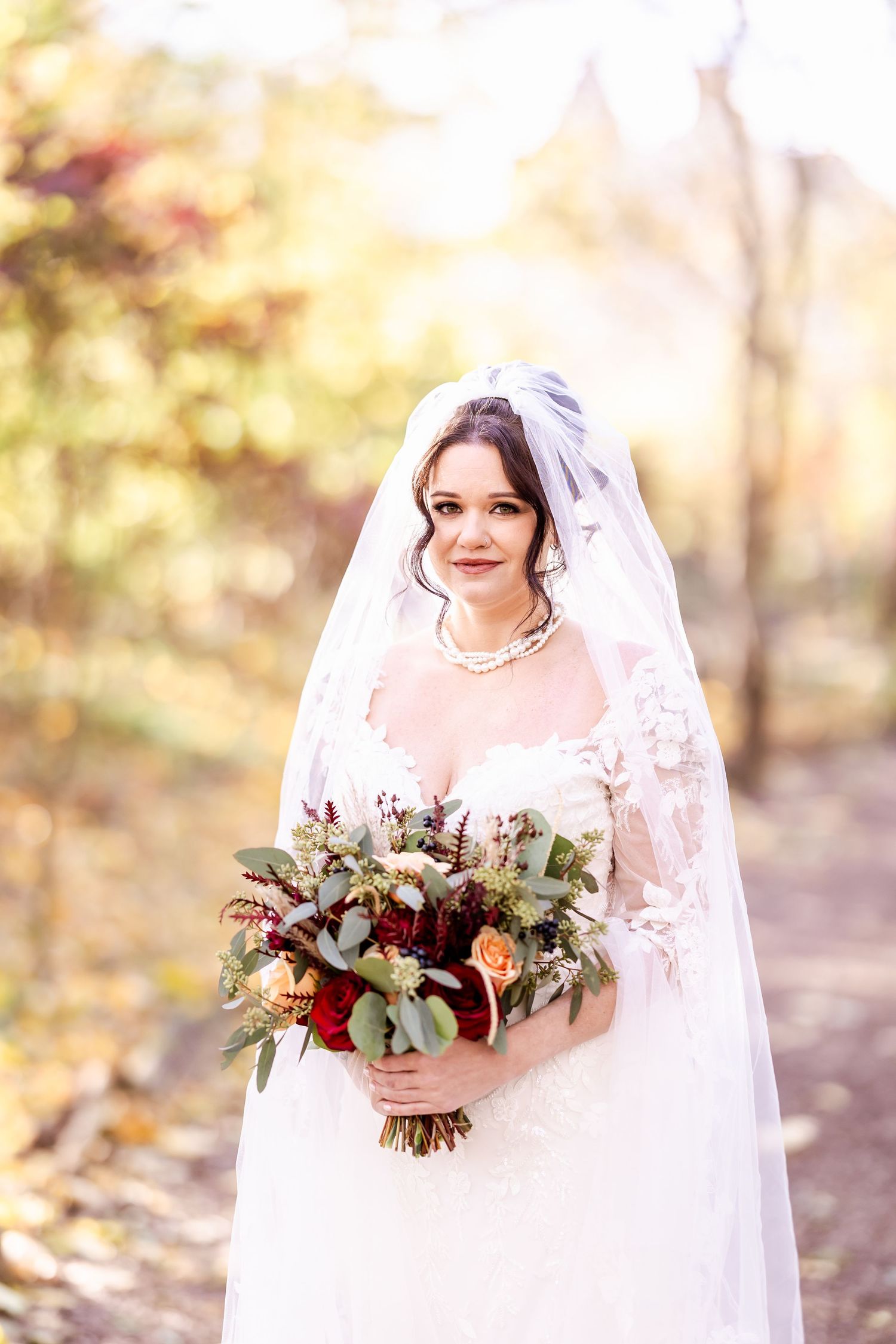 A bride in a white lace gown holds an autumn-toned bouquet during a fall outdoor wedding photoshoot.