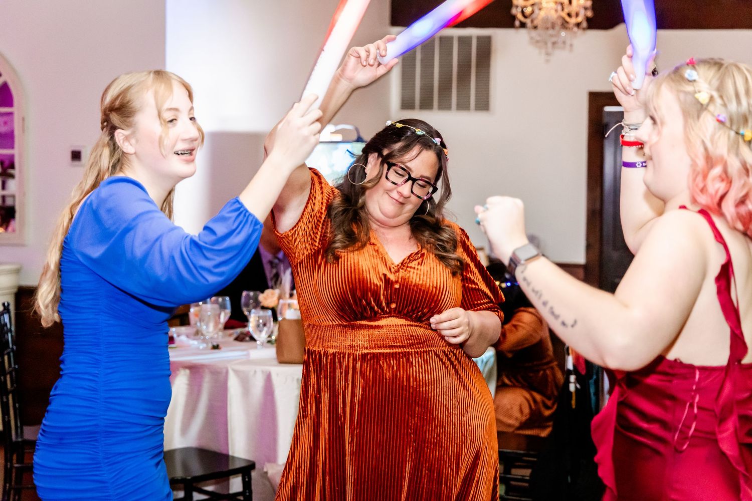 Wedding party in jewel-toned dresses dancing and celebrating at an indoor reception with purple lighting.