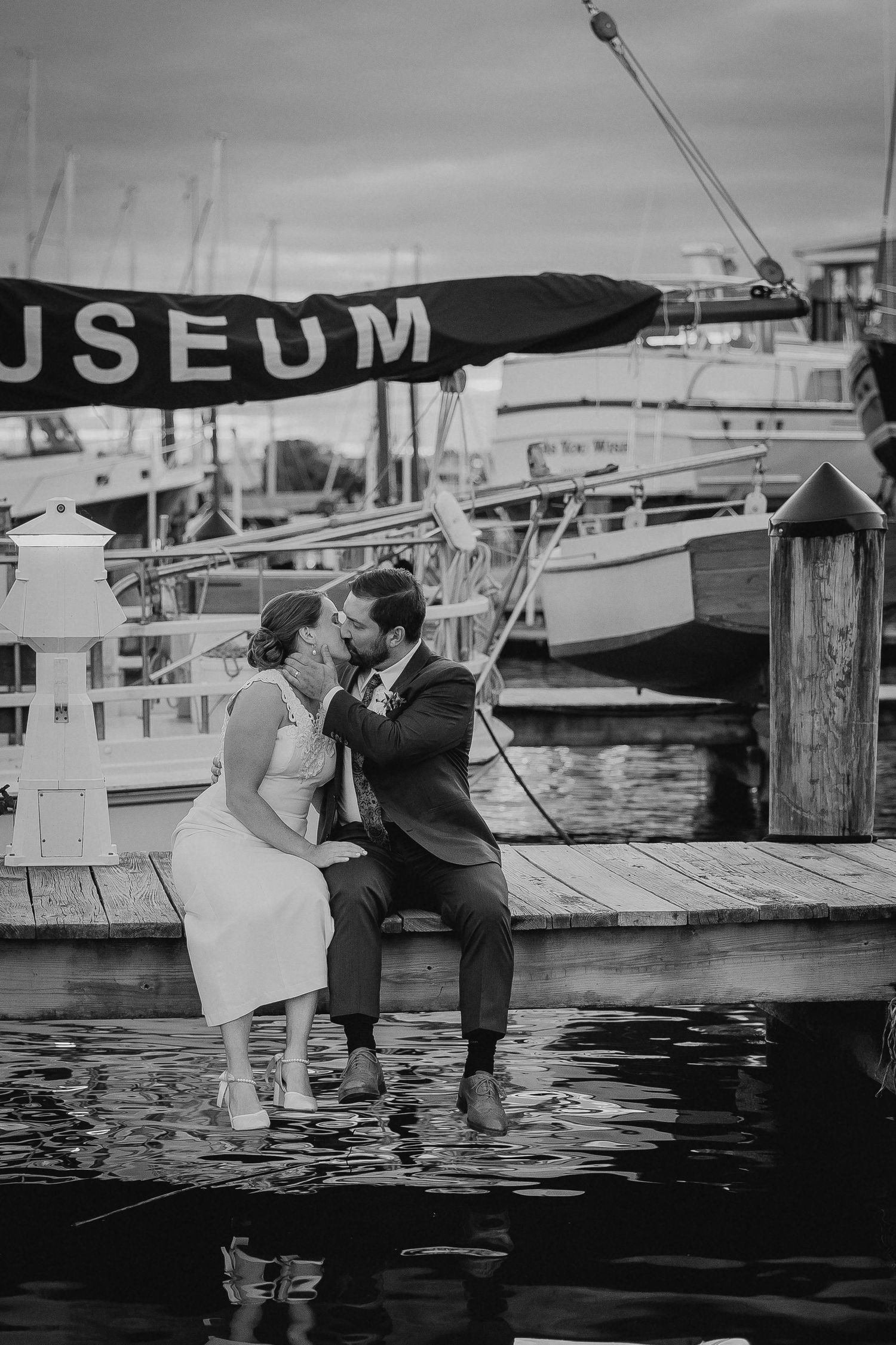 Black and white photo of a couple kissing on a dock near a museum and sailboats on their wedding day.