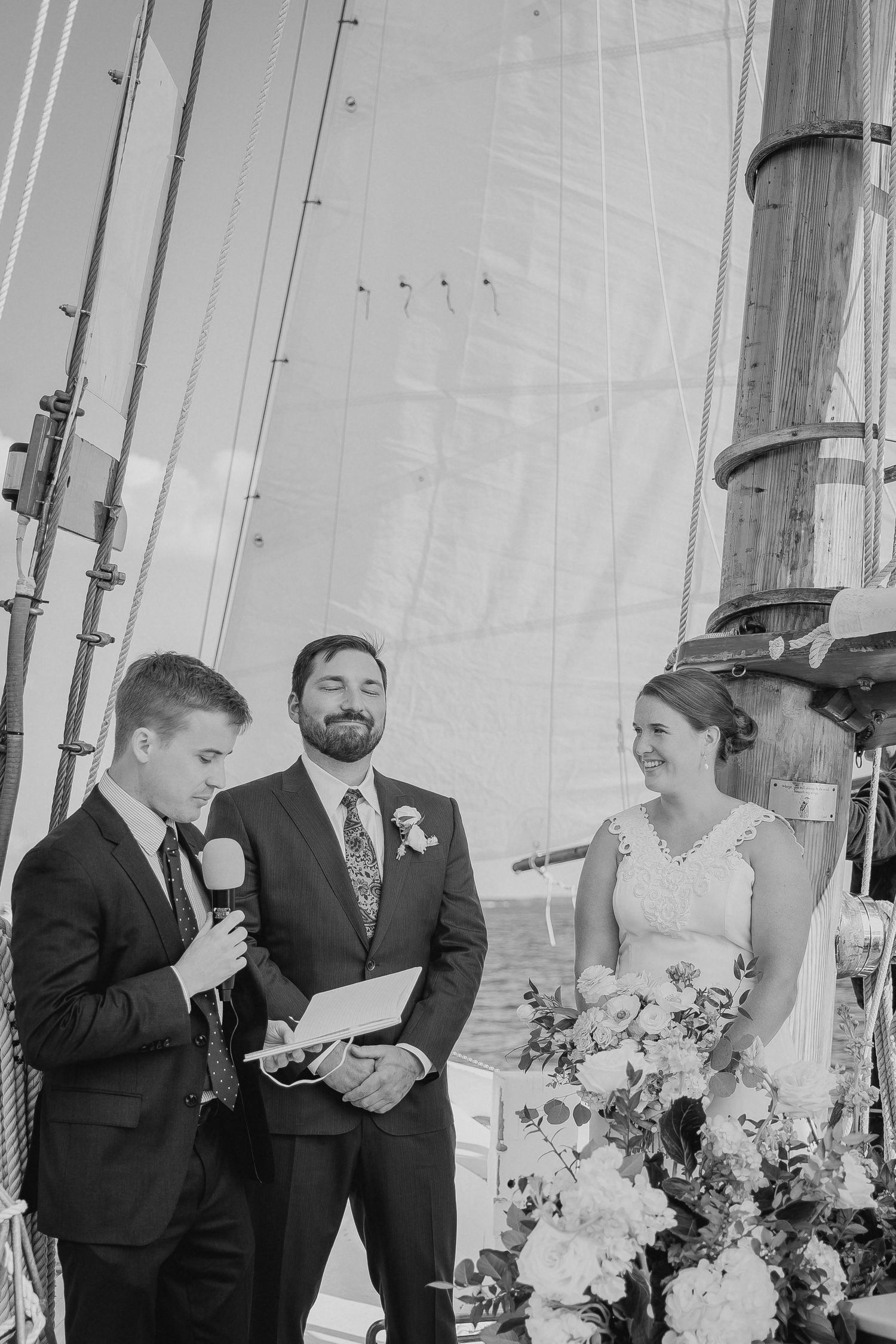 Black and white photo of a wedding ceremony on a sailboat with bride, groom, and officiant holding microphone.