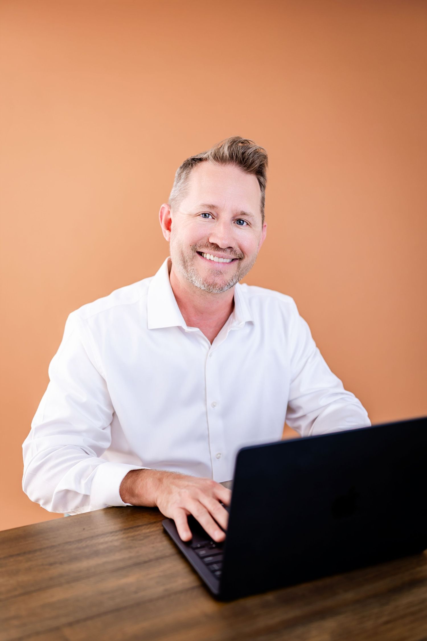 Smiling man in white shirt working on laptop at wooden desk against orange background.