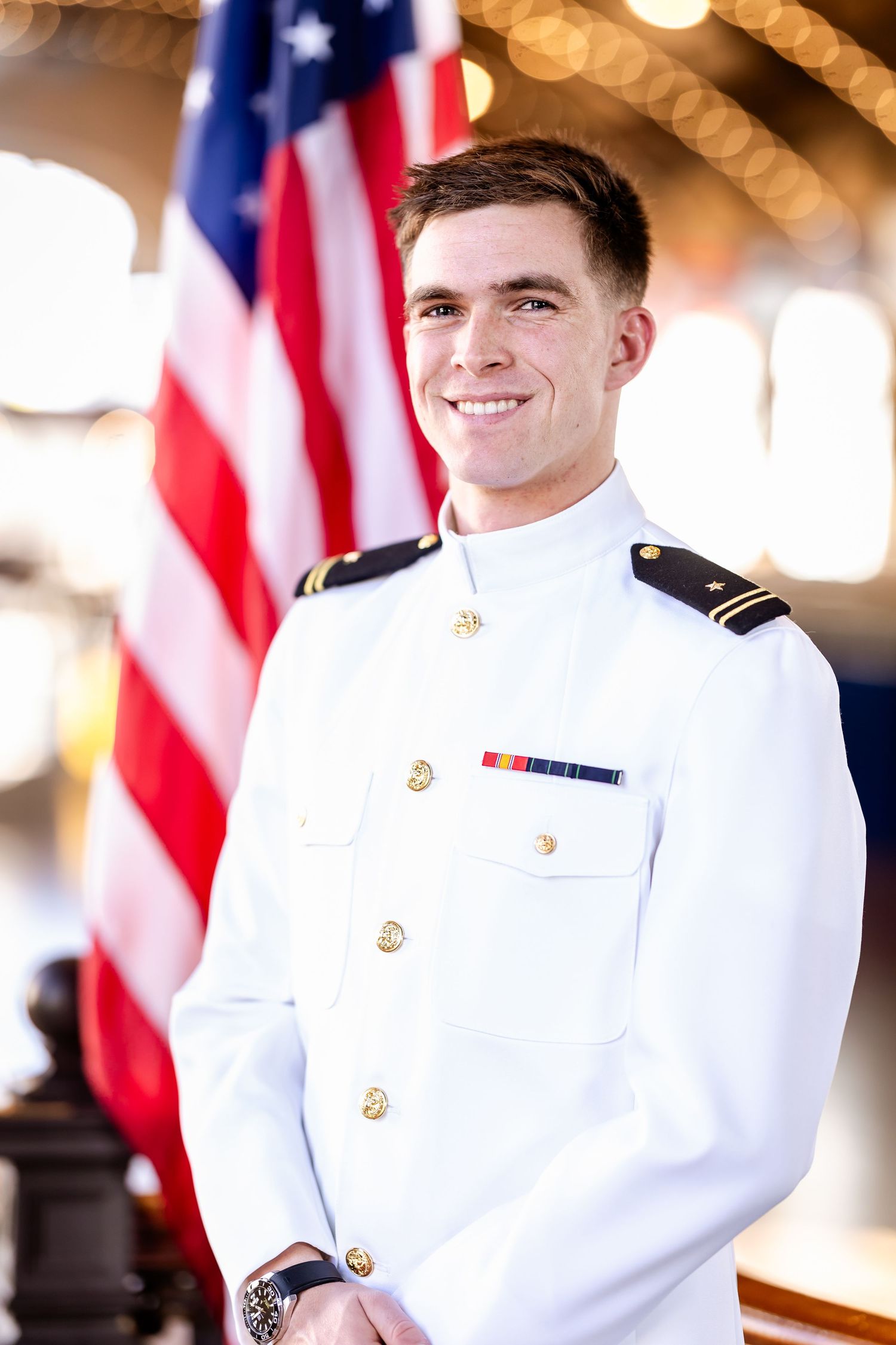 Young man in white naval dress uniform smiling before an American flag.