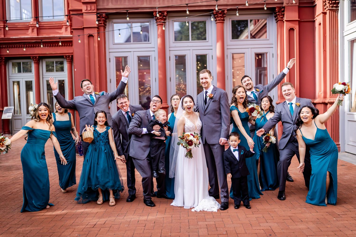 Wedding party in teal blue dresses and suits poses playfully on red brick steps for group photos.