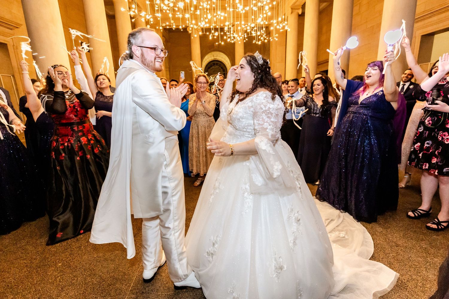 Wedding couple dancing in a grand ballroom surrounded by cheering guests and ornate columns.