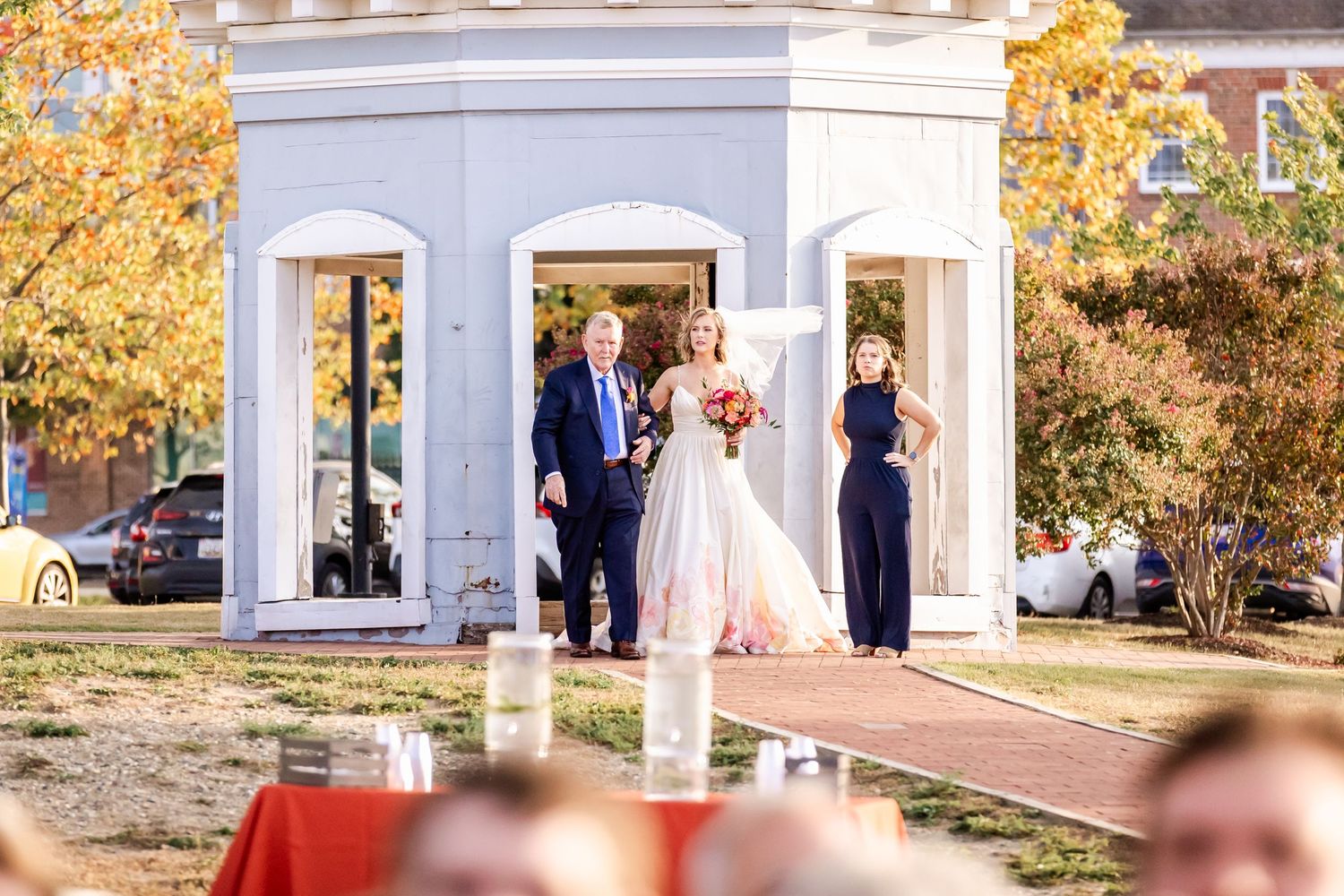 A wedding ceremony takes place under an elegant white gazebo surrounded by autumn foliage.