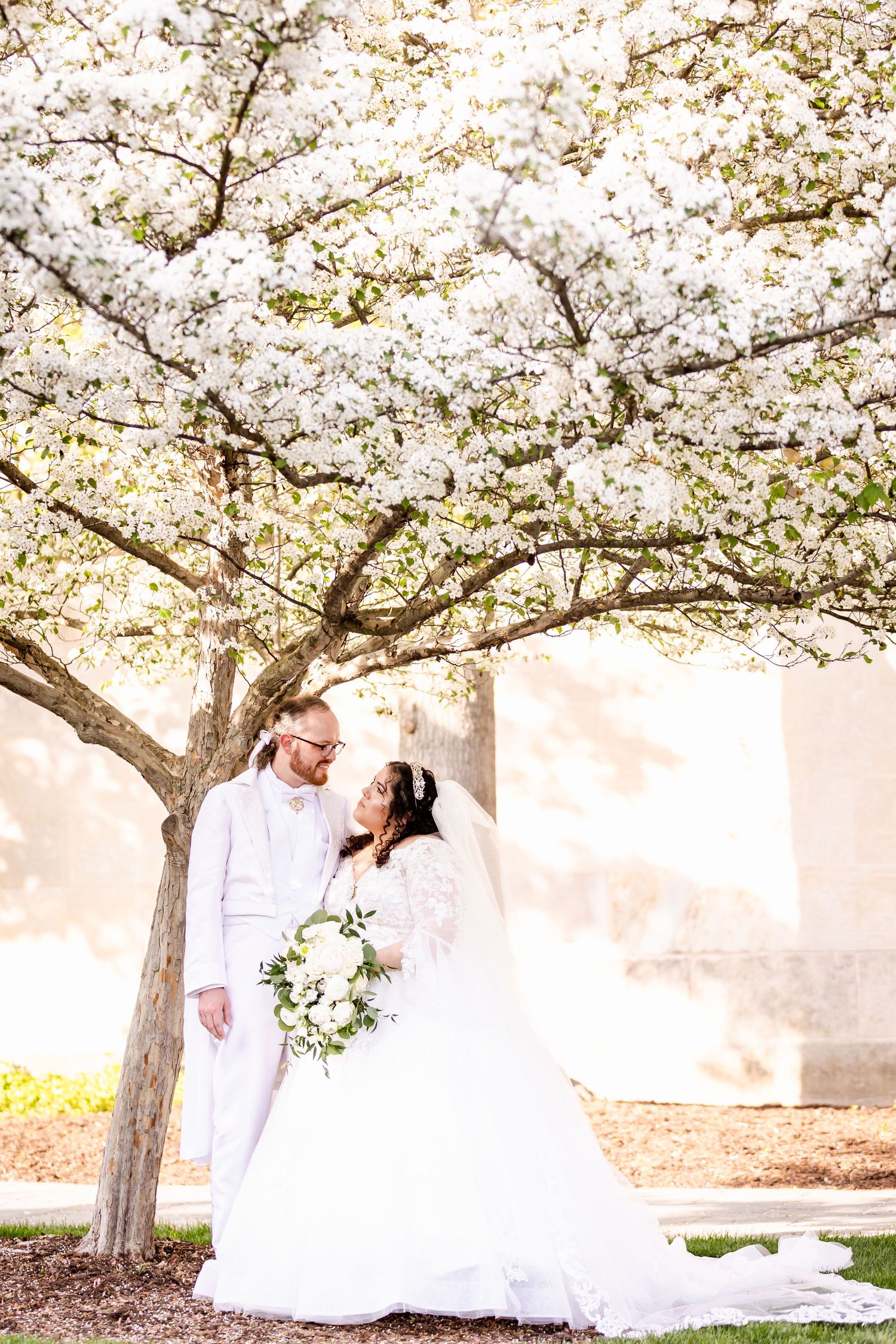 Couple poses beneath blooming cherry blossom tree in elegant white wedding attire.