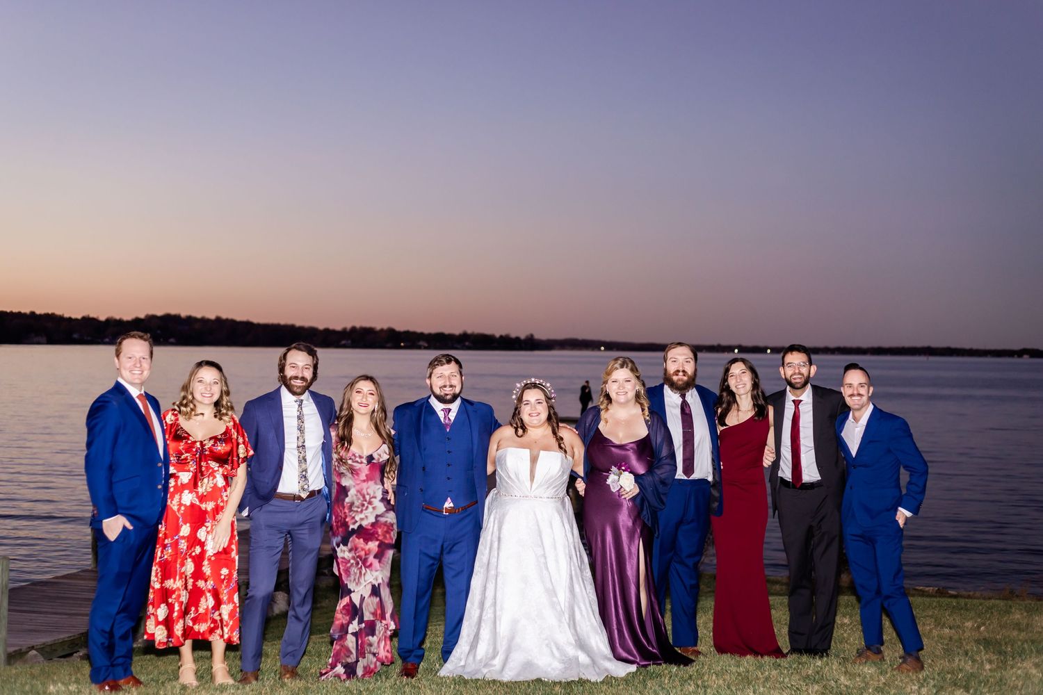 Wedding party poses for group photo at waterfront during sunset.