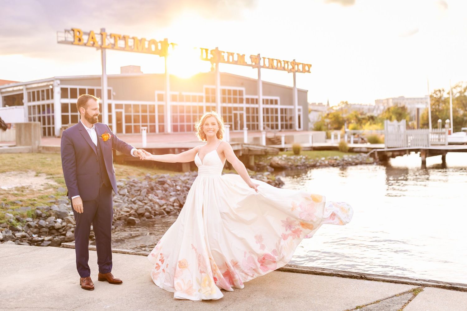 A romantic sunset wedding photo on a waterfront boardwalk with illuminated building signage in the background.