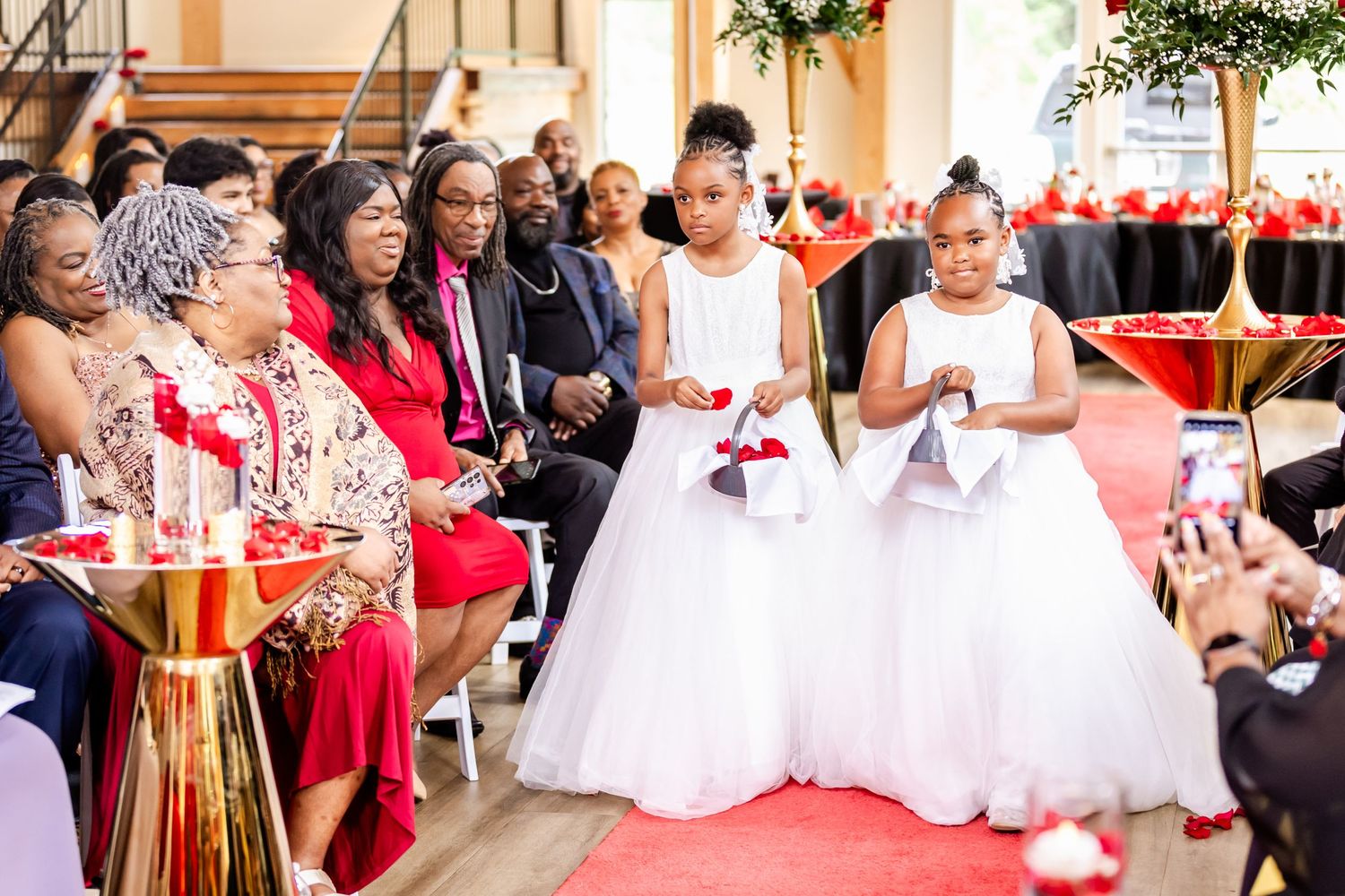 Flower girls in white dresses walk down the red carpet aisle during an elegant ceremony.