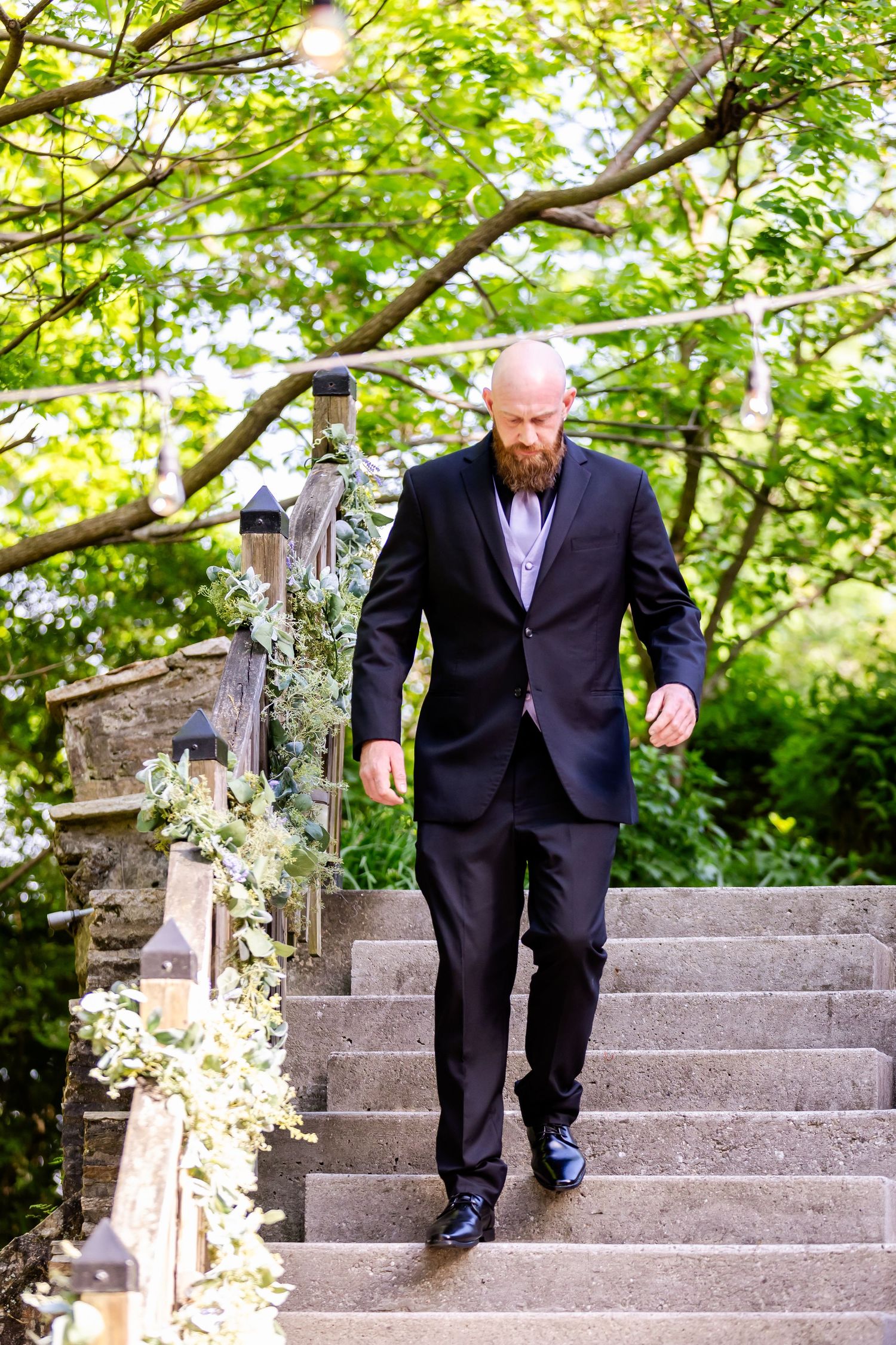 Person in dark suit descends stone staircase decorated with floral arrangements in garden setting.