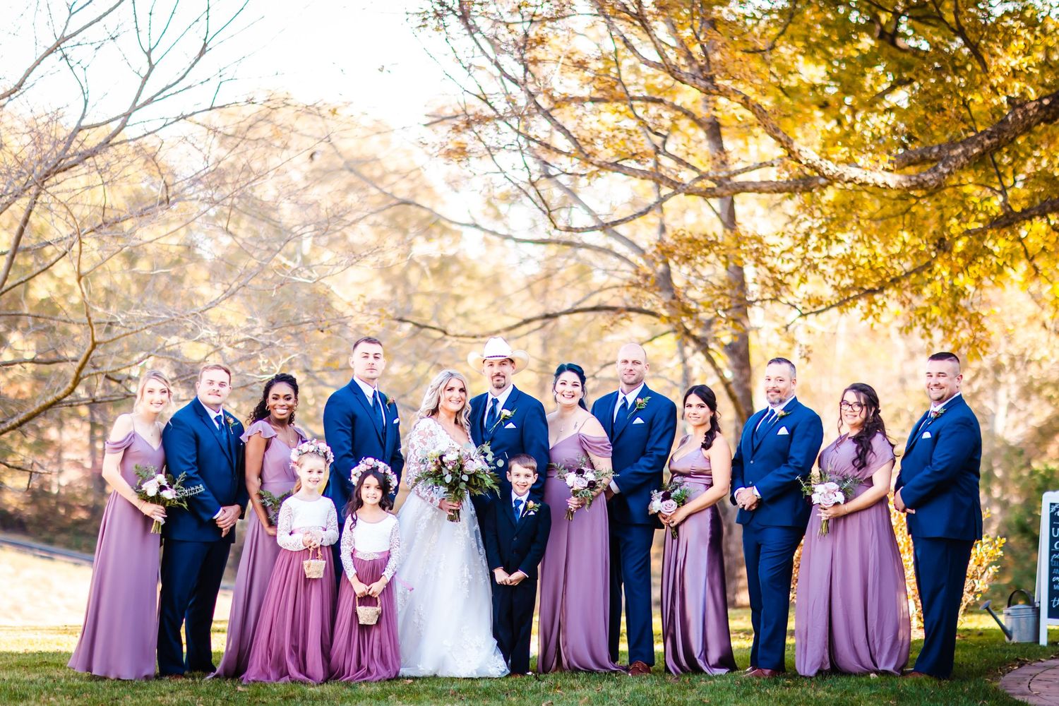 Wedding party in navy suits and mauve bridesmaid dresses poses together on sunny autumn day with golden trees.