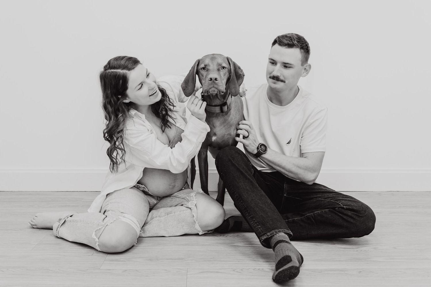 A happy couple sits on the floor playing with their Dachshund dog in a black and white photograph.