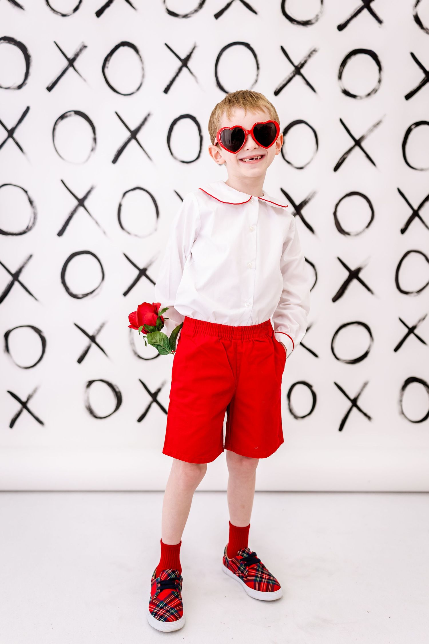 Stylish young figure in red shorts and sunglasses poses with roses against X's and O's patterned background.