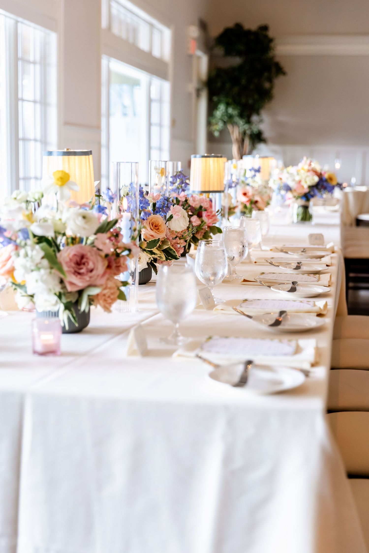 Long wedding reception table with delicate floral centerpieces and white linens in bright sunlit room.