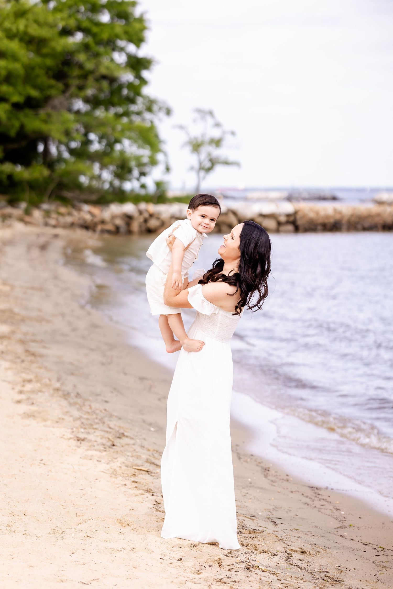 A tender beach portrait with a person in white holding a small child against a coastal backdrop.