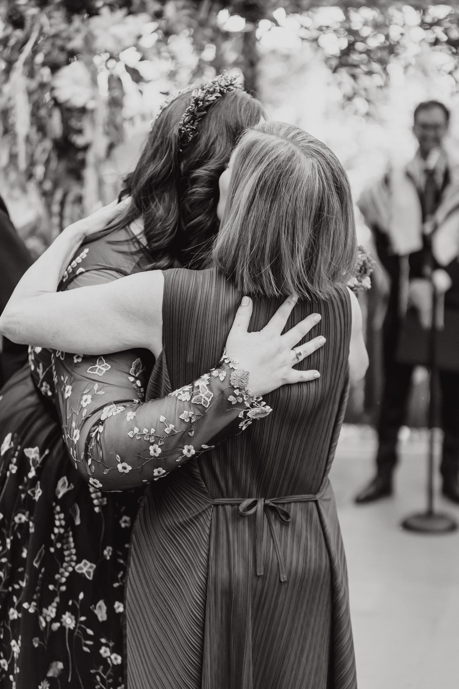 Two women in floral and striped dresses share an emotional embrace at a formal event.
