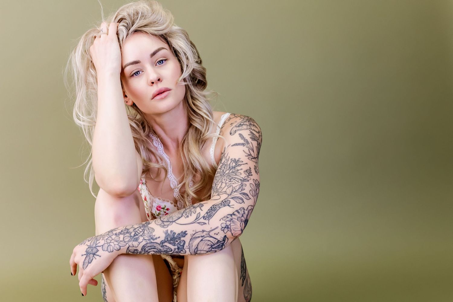 Fashion portrait with artistic tattoo sleeves and blonde hair against olive green background.