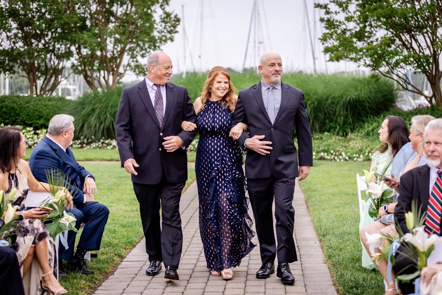 Guests in formal attire mingle on a garden path during an outdoor wedding reception.