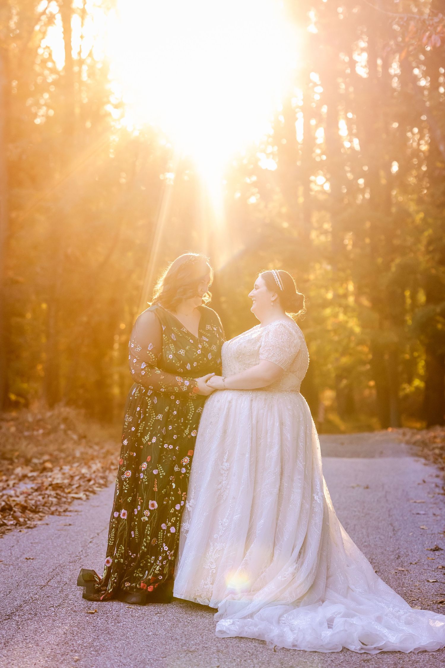 Two brides sharing intimate moment in sunset forest with white wedding dress trailing behind.
