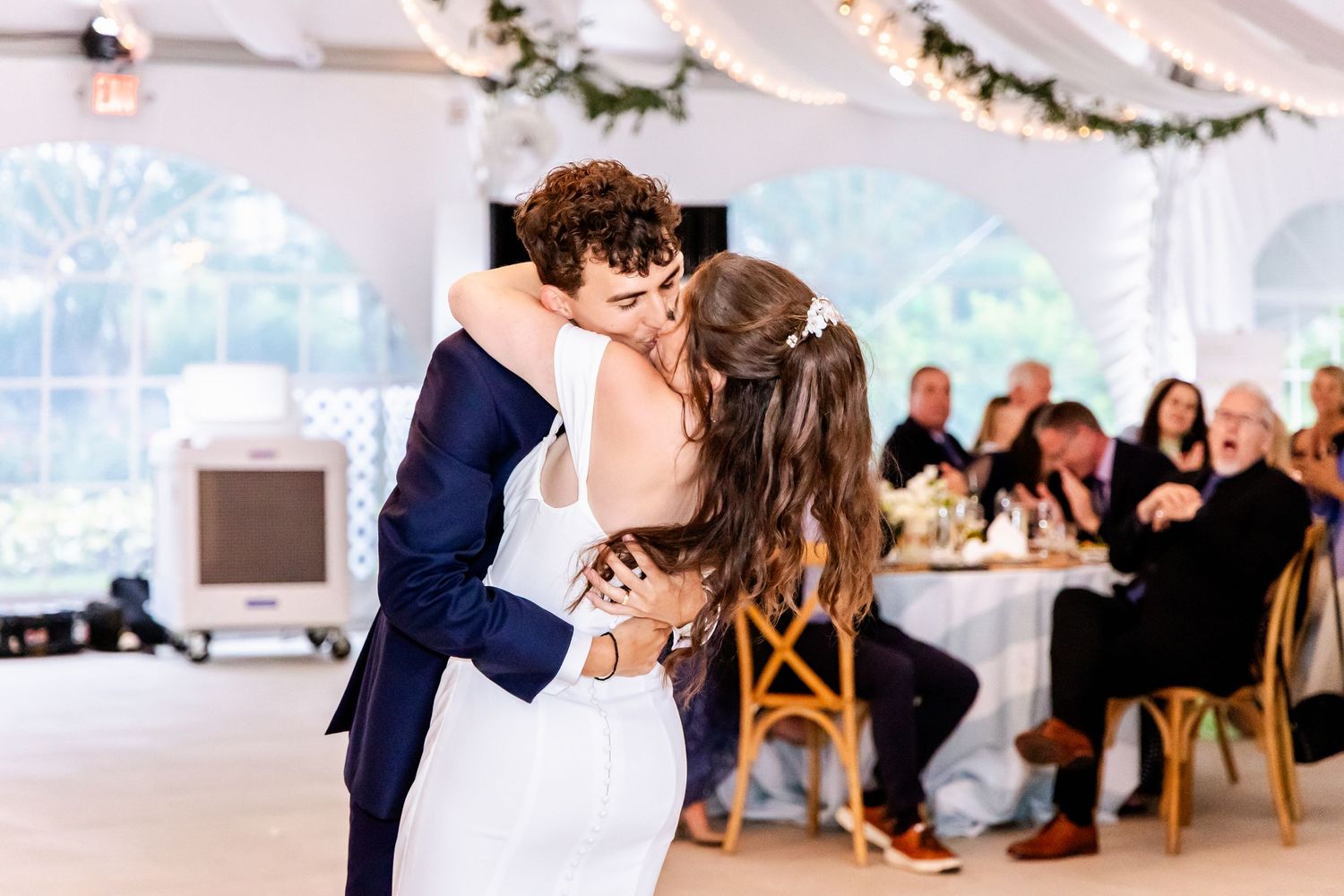 Wedding reception venue with elegant white decor and guests seated at long tables under string lights.