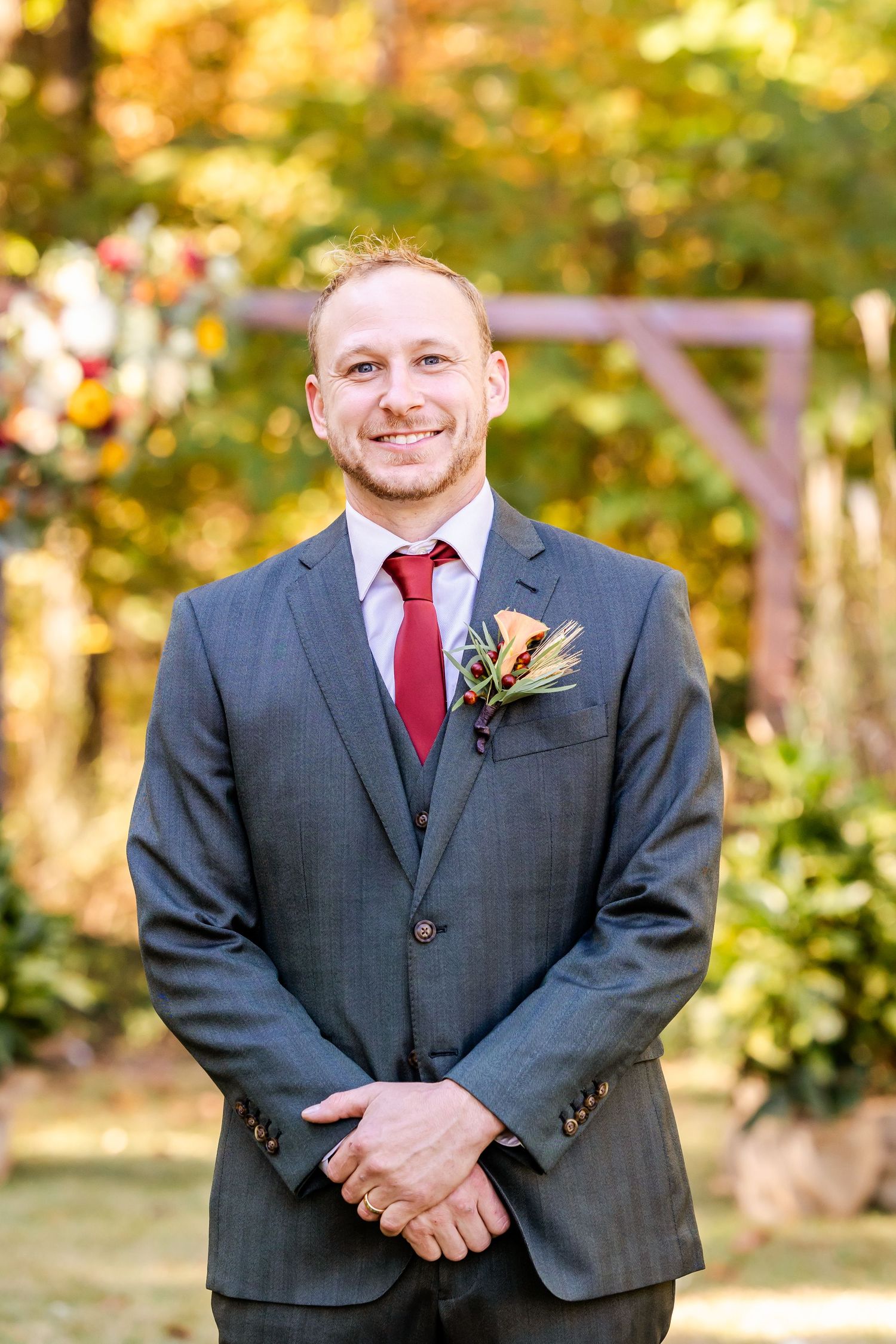 Groom in charcoal suit with red tie poses outdoors during fall wedding.