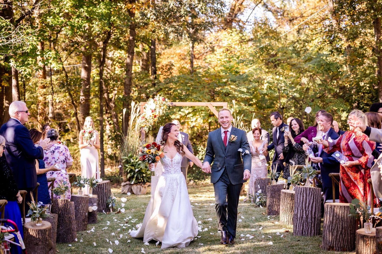 Newlyweds walk down a tree-lined aisle with flower petals while wedding guests celebrate their exit.