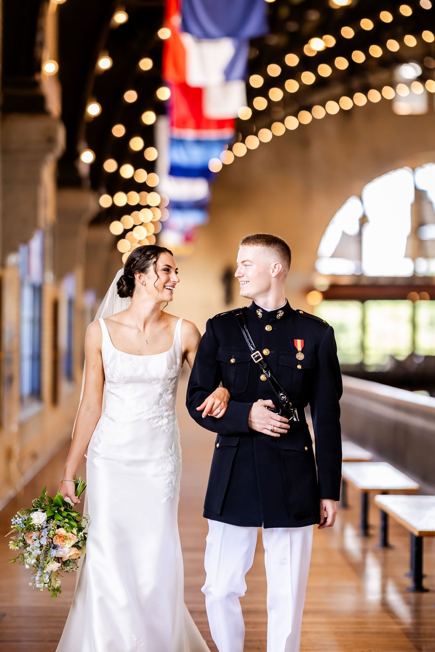 A wedding couple walking together under string lights with military dress uniform and white bridal gown.
