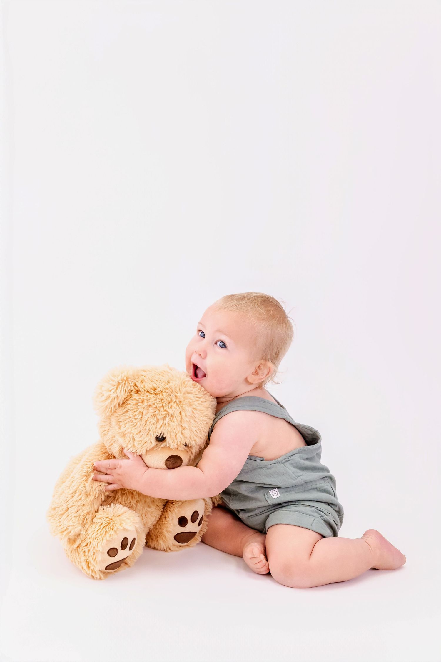 Baby in gray romper cuddling with soft brown teddy bear on white backdrop.