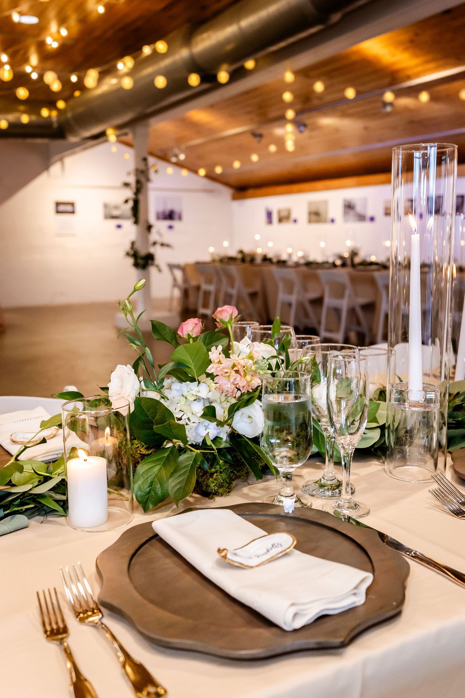 Elegant wedding reception table with floral centerpiece, gold charger plates, crystal glassware, and string lights in a rustic venue.