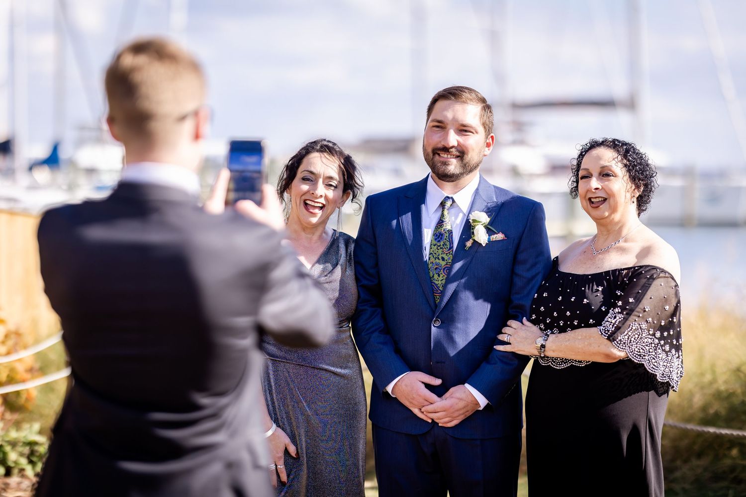 Groom smiling with two women at waterfront wedding ceremony while guest photographs them on smartphone.