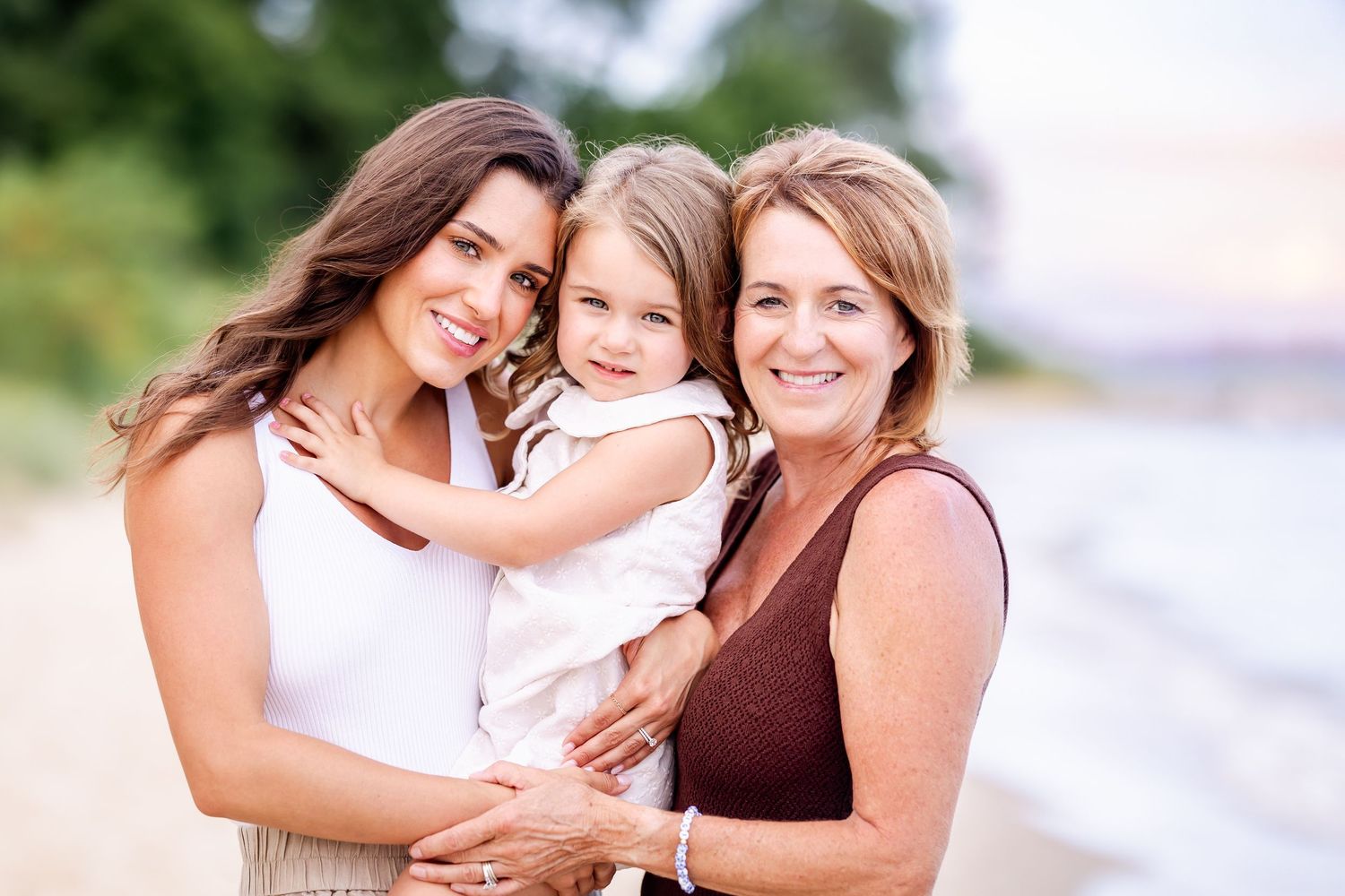 Three people share warm embraces and smiles while posing together on a sunny beach in coordinated outfits.
