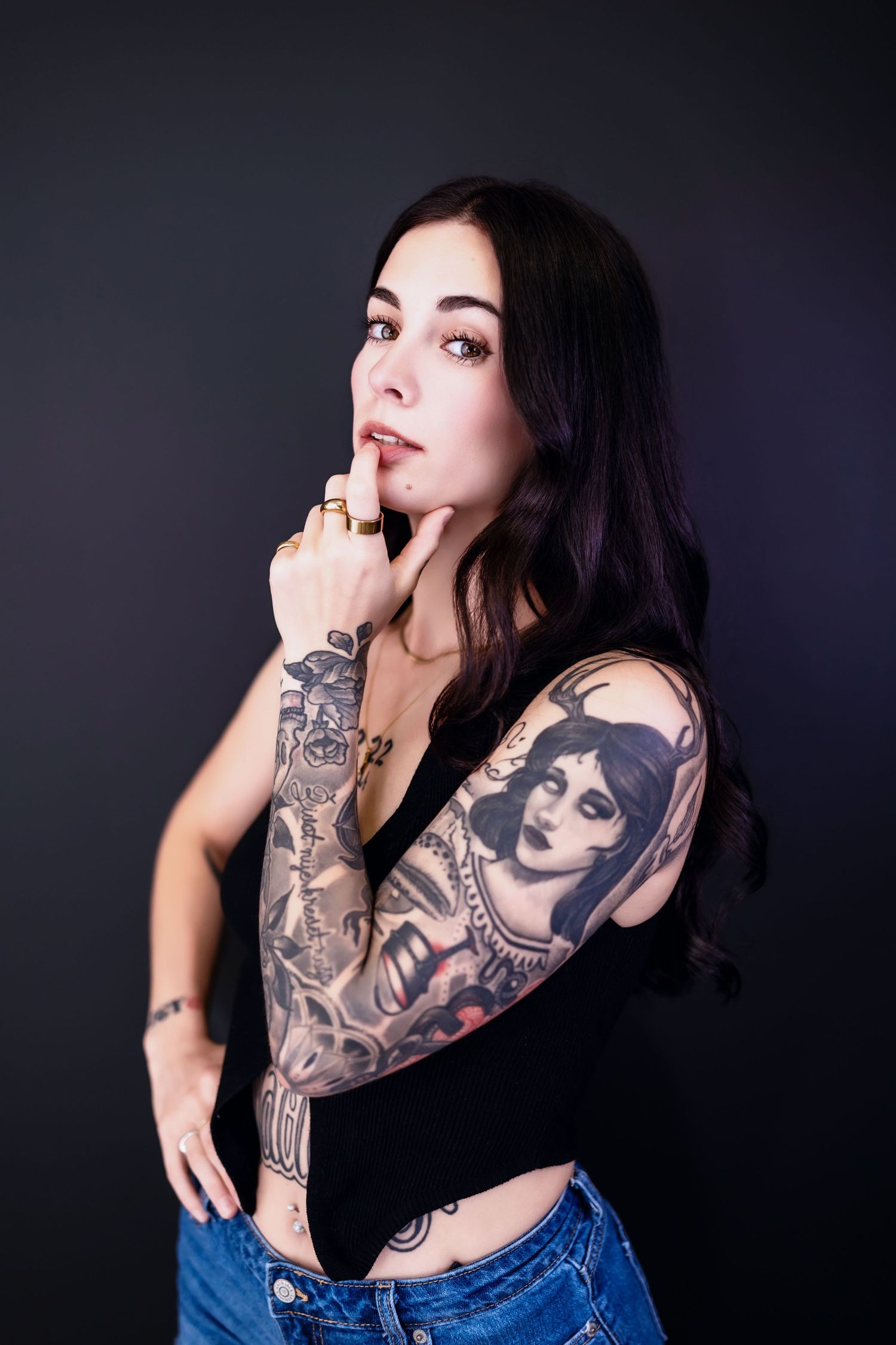 Young woman with long dark hair and full sleeve tattoos poses in black crop top and jeans against dark background.