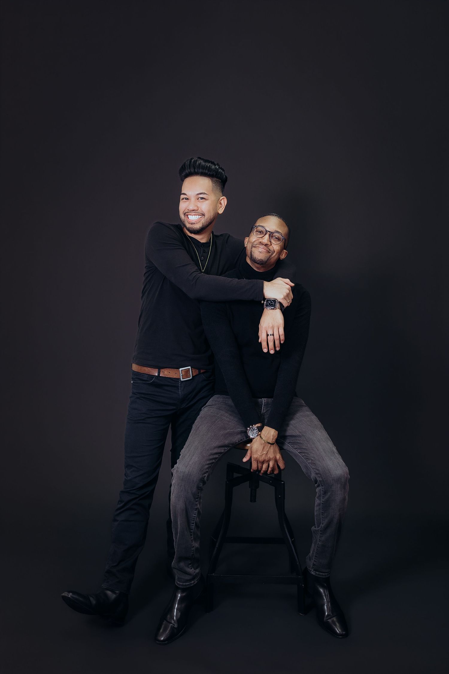 Two men pose playfully in dark outfits against a black backdrop in a professional studio setting.
