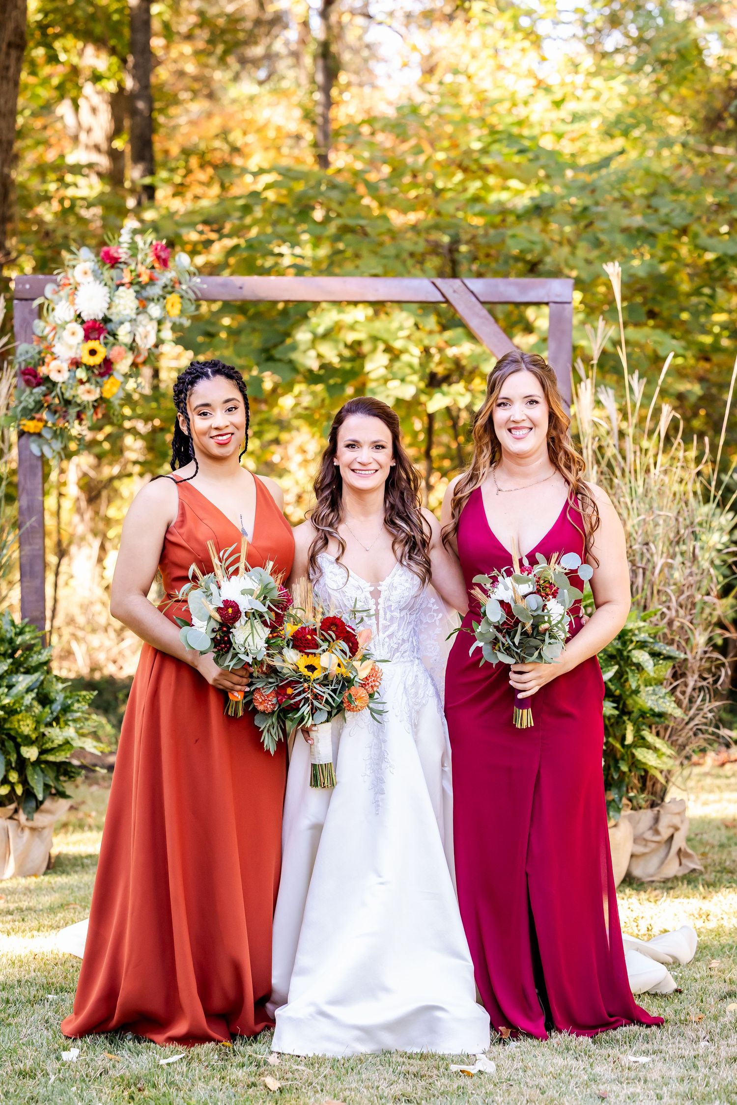 Bridal party in burgundy and rust-colored dresses poses outdoors with autumn scenery.